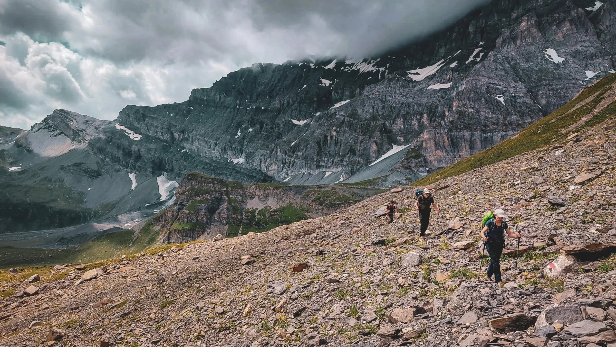 Randonneurs sur un sentier alpin, entourés de montagne majestueuse et de nuages dramatiques.