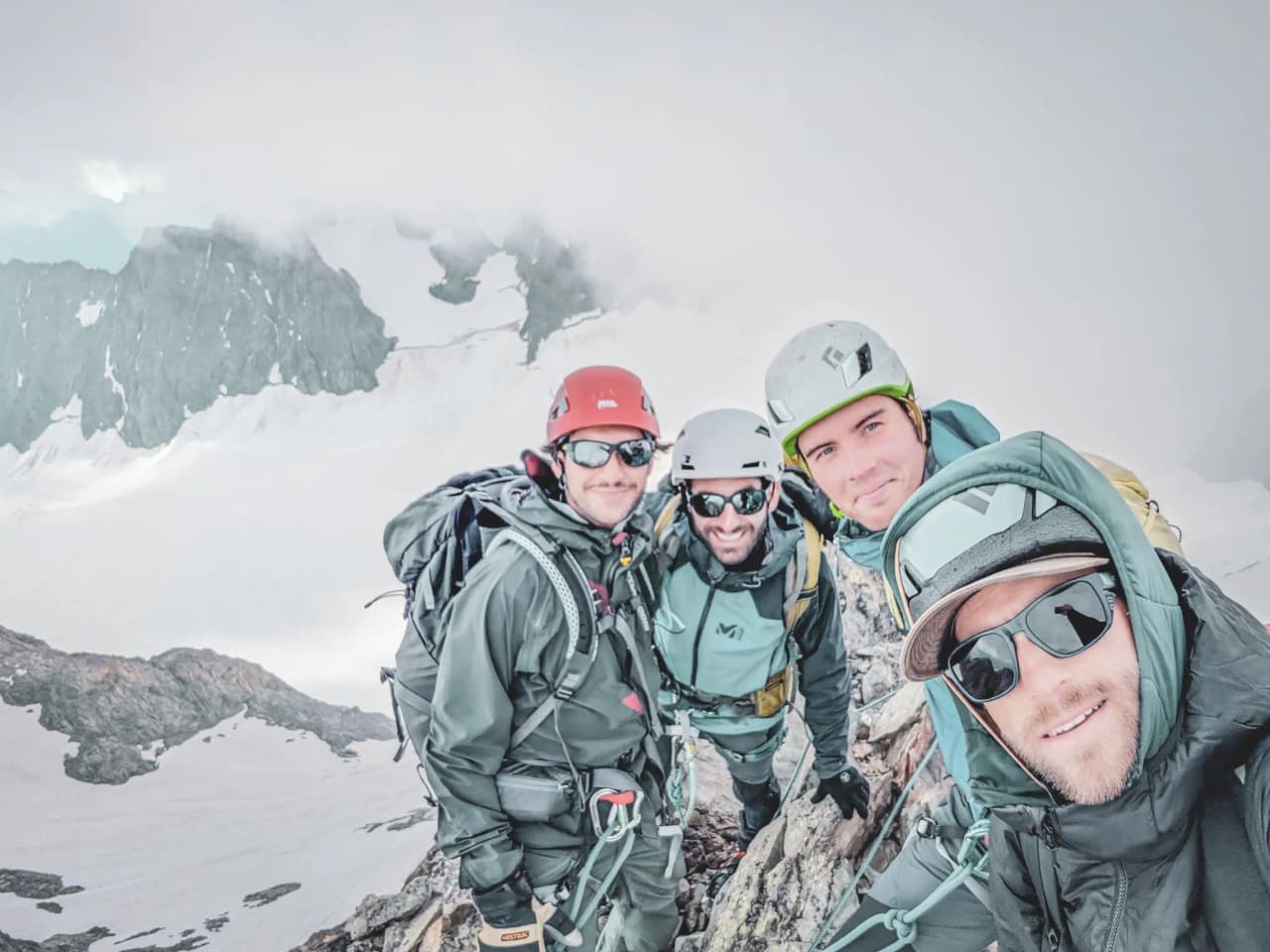Four smiling climbers at the summit, with the majestic scenery of the Écrins in the background.