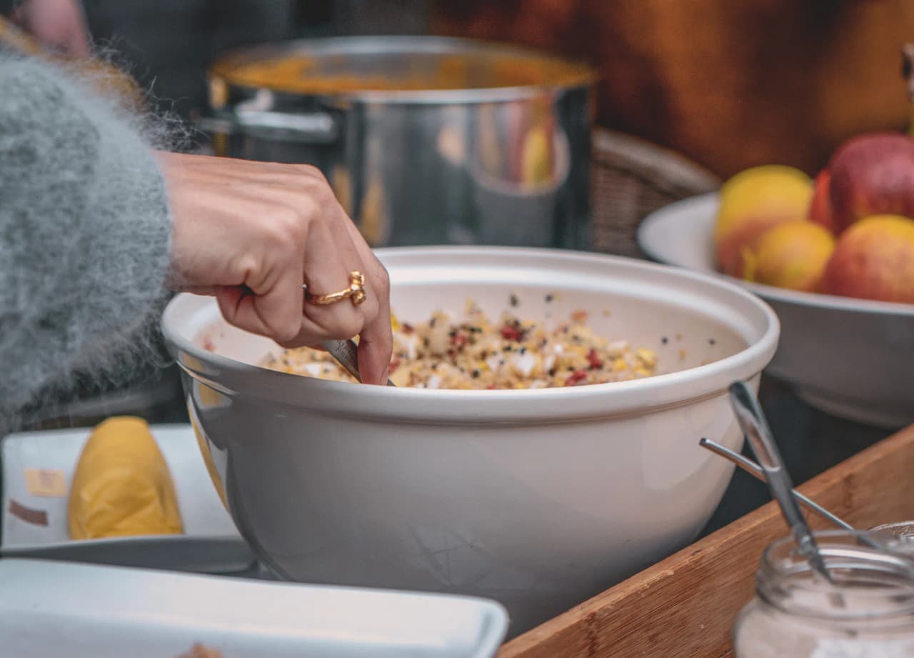 Een hand met een gouden ring mengt ingrediënten in een grote witte kom, gevuld met wat een kleurrijk mengsel van granen lijkt te zijn. Op de achtergrond staat een metalen steelpan met daarin een