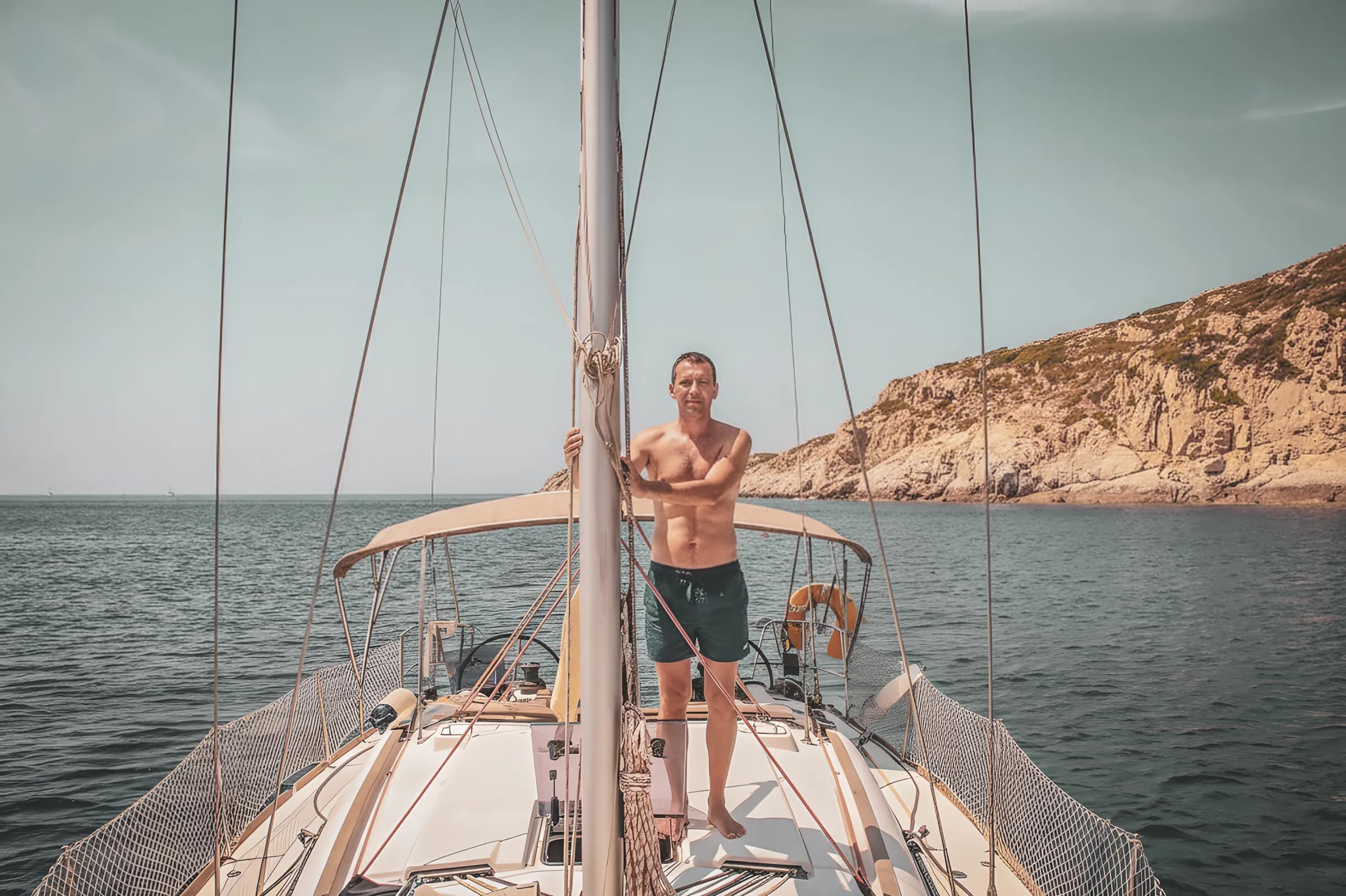 Un homme souriant en maillot, sur un voilier face à la magnifique côte rocheuse de Corse.