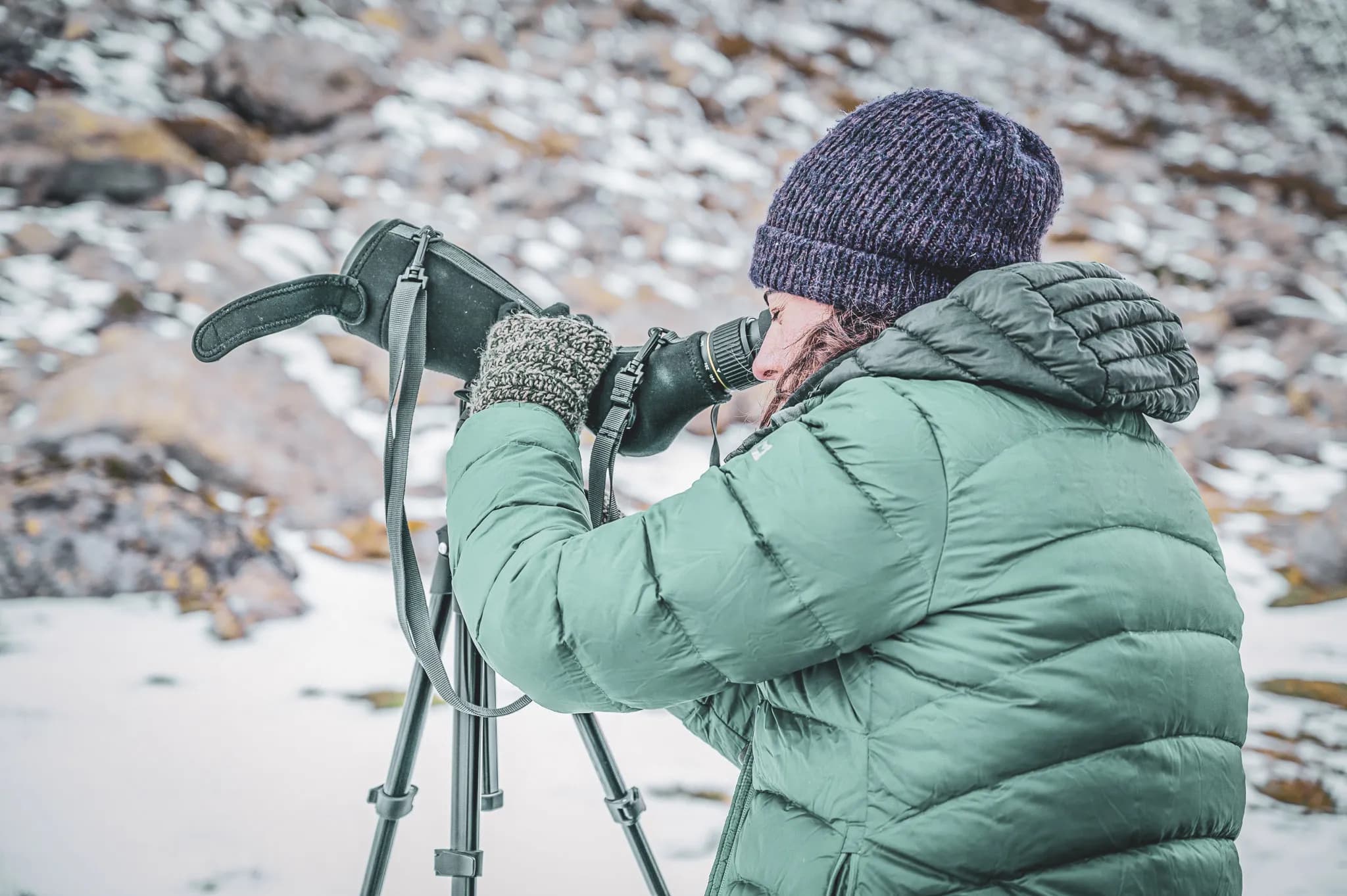A hiker in a green down jacket observes wildlife through a telescope on a snowy path.