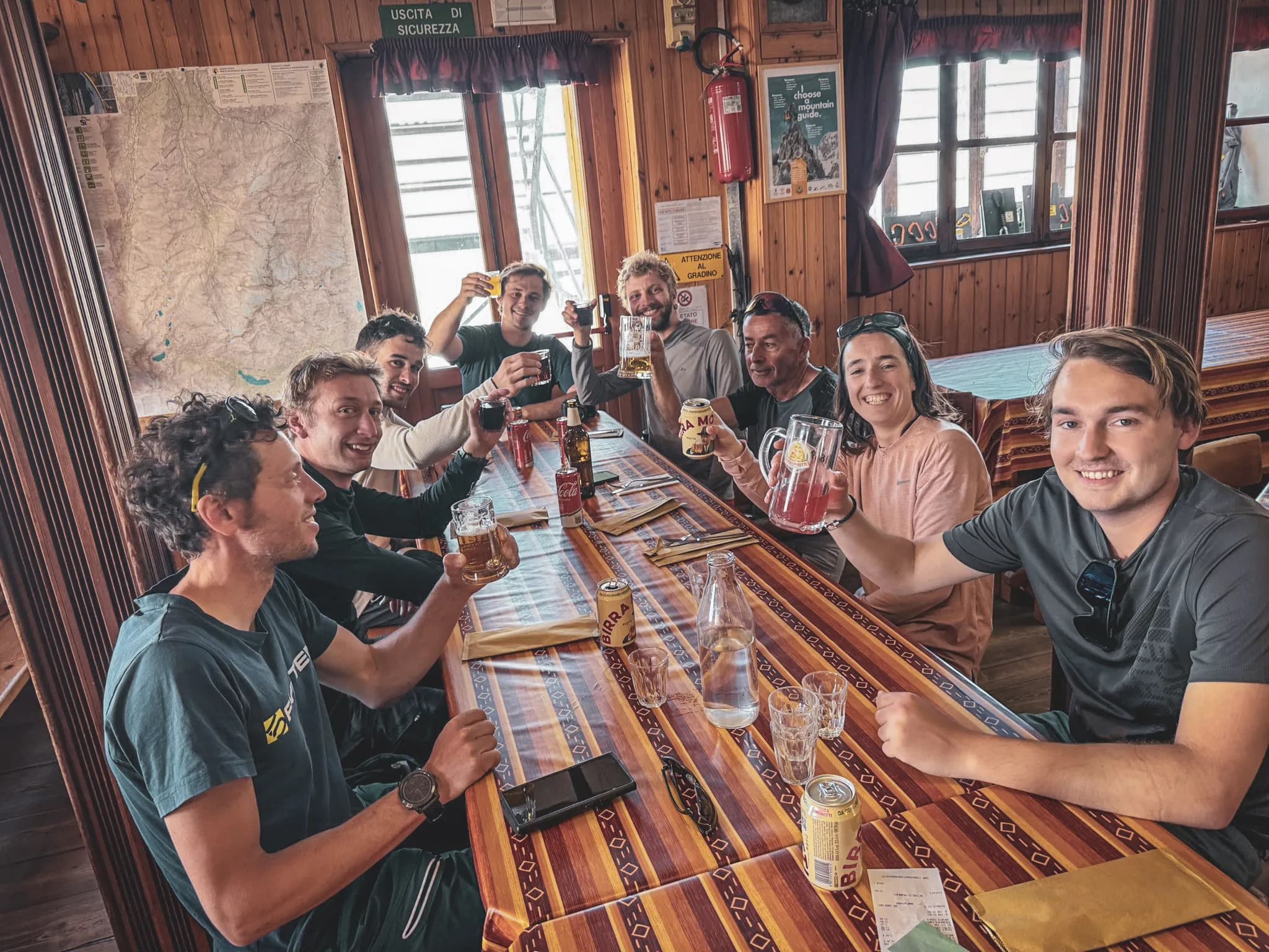 A group of climbers celebrate their ascent to the summit, drinks in hand, in a cosy Mountain hut.