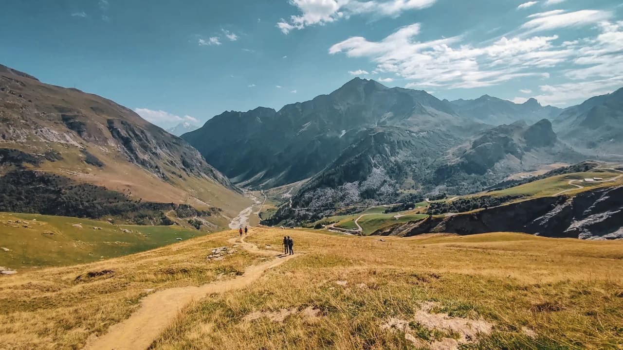 Two hikers on a green path, surrounded by majestic Alpine peaks under a blue sky.