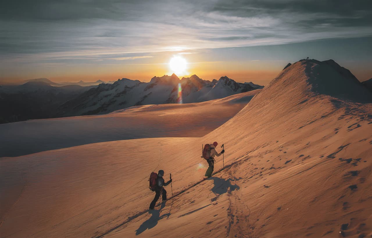 Deux skieurs gravissent une pente ensoleillée, entourés de montagnes majestueuses au lever du soleil.
