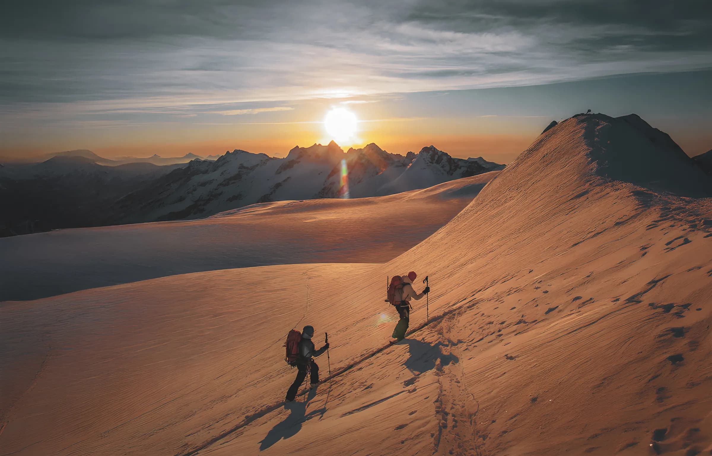 Deux skieurs gravissent une pente ensoleillée, entourés de montagnes majestueuses au lever du soleil.