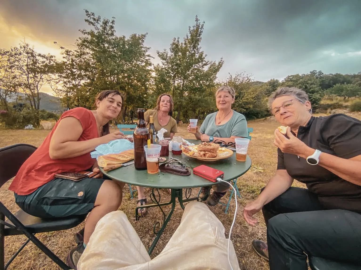 Vier lachende vrouwen rond een tafel, die een maaltijd delen op het platteland bij zonsondergang.
