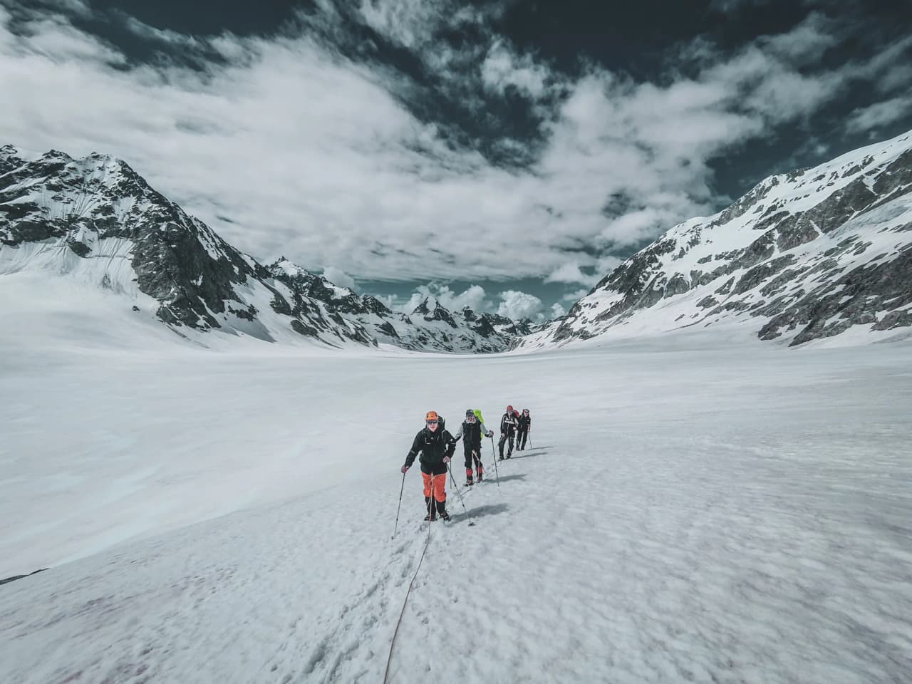 Five hikers make their way across a vast glacier, surrounded by majestic Alpine mountains.