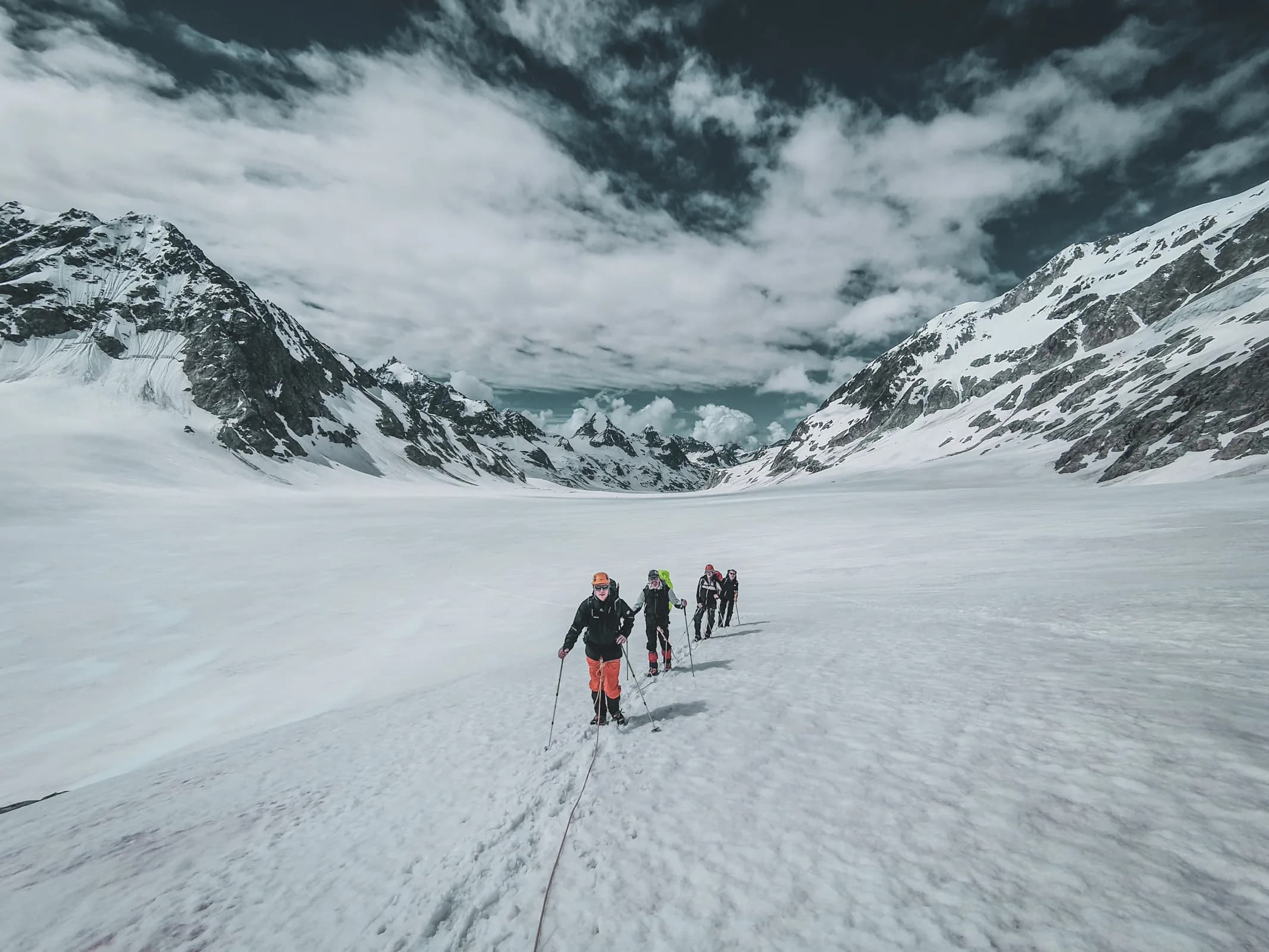 Five hikers make their way across a vast glacier, surrounded by majestic Alpine mountains.