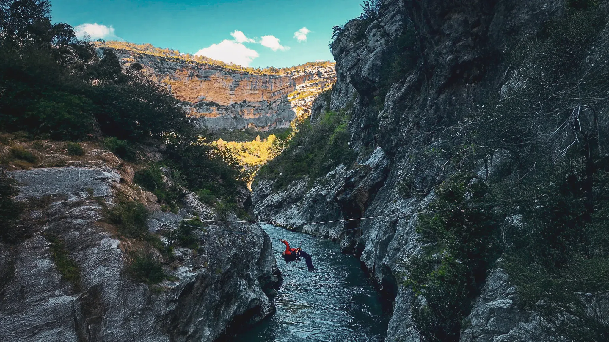 Un grimpeur en rouge traverse une rivière sur un câble, entouré de falaises majestueuses.