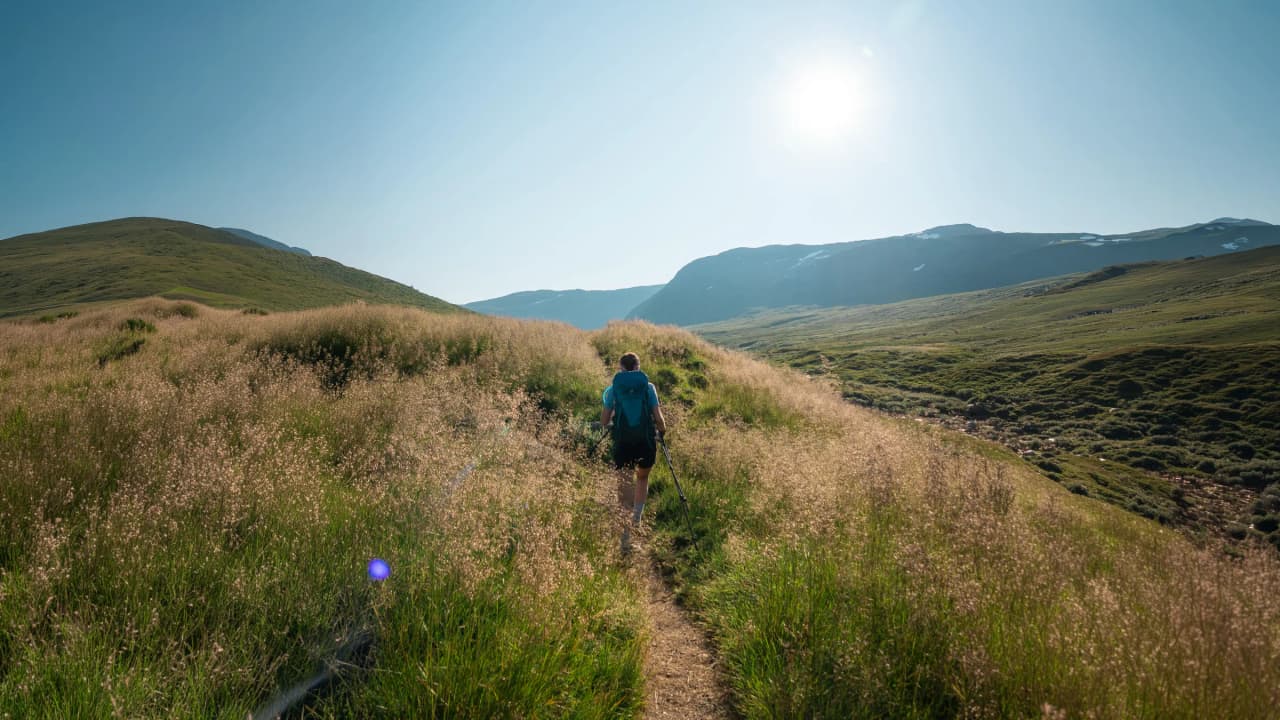 A solitary hiker makes his way along a green path, surrounded by mountains in brilliant sunshine.