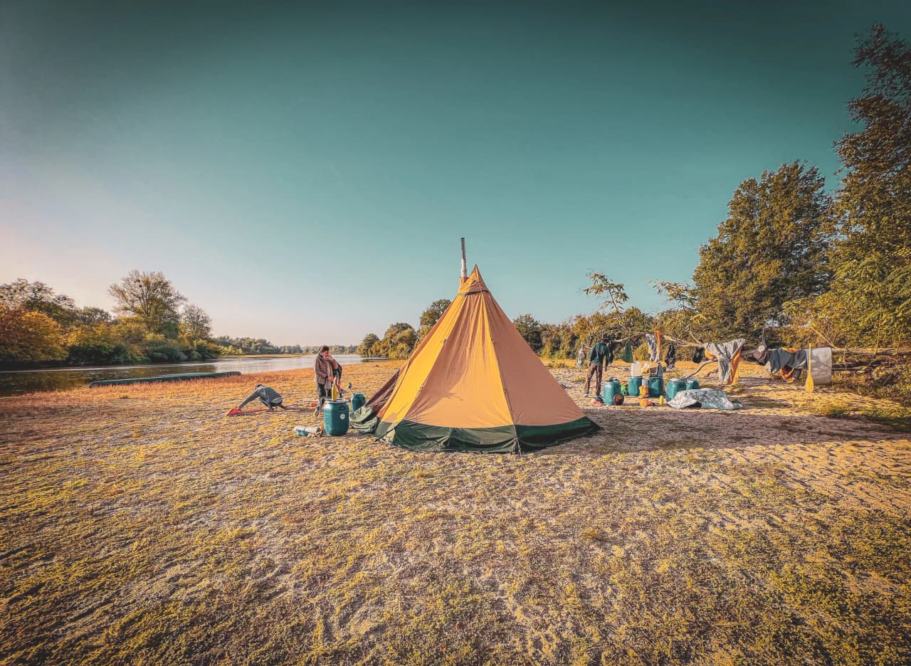 Een kamp aan de rivier met een grote oranje kegelvormige tent. Enkele mensen zijn zichtbaar, één hanteert apparatuur bij de tent, terwijl anderen zich eromheen druk maken. Van