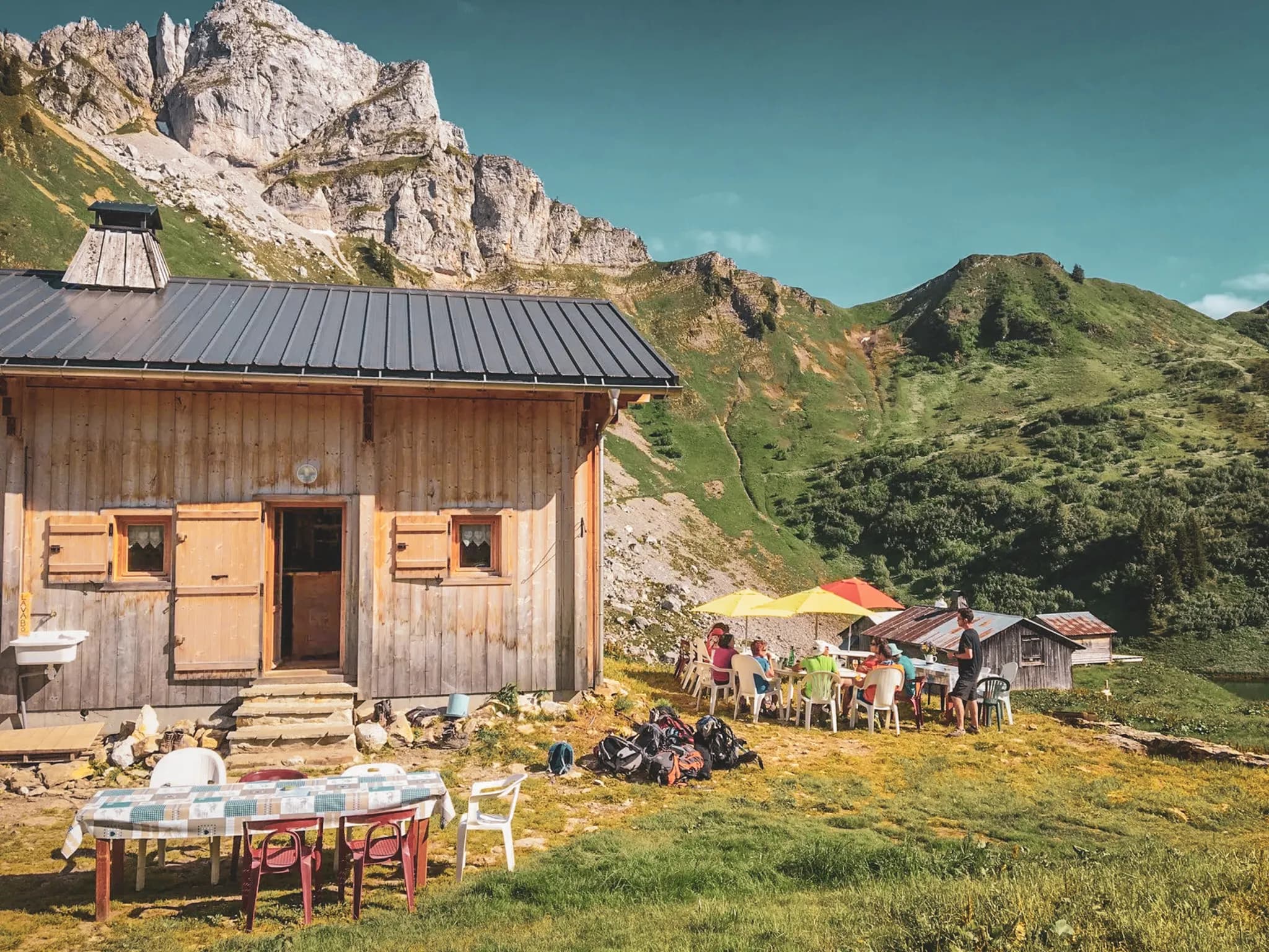 Refuge alpin ensoleillé, randonneurs réunis autour de tables colorées, entourés de montagnes verdoyantes.