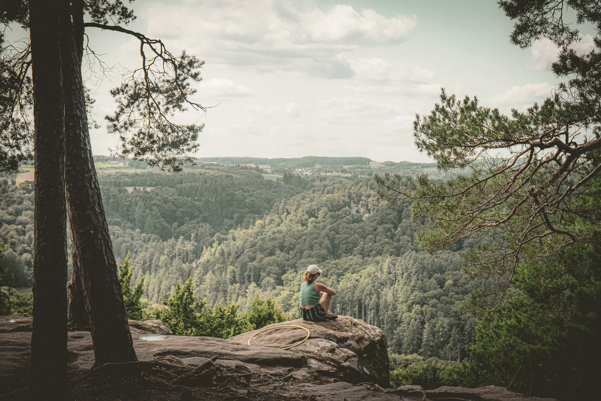 jeune personne assise sur un rocher, contemplant des forêts verdoyantes sous un ciel nuageux.