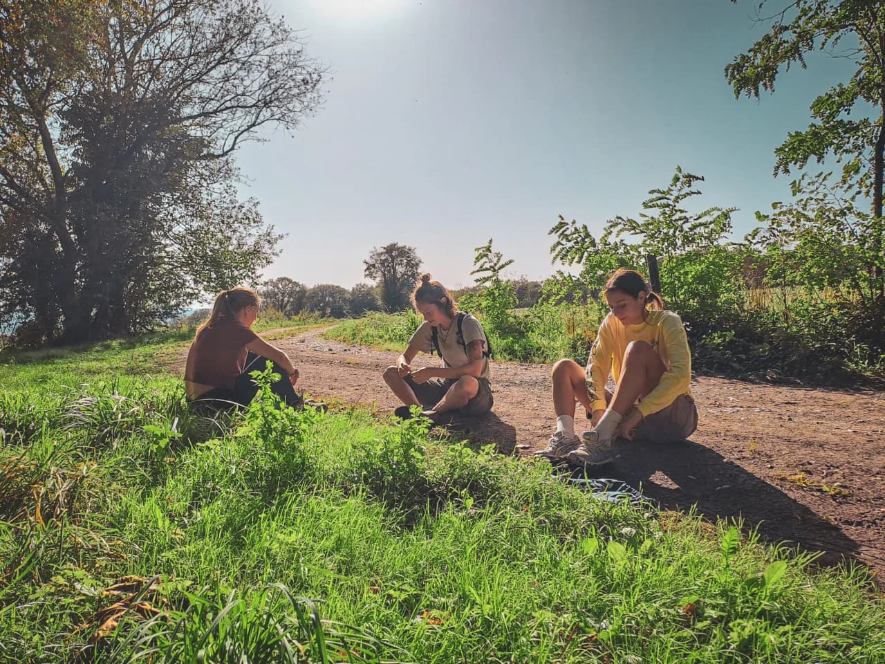 Drie mensen zitten op een zandpad, omgeven door groen. Twee van hen leunen naar de grond en hebben een ontspannen moment, terwijl de derde, met een geel t-shirt aan, wacht tot de zon opkomt.