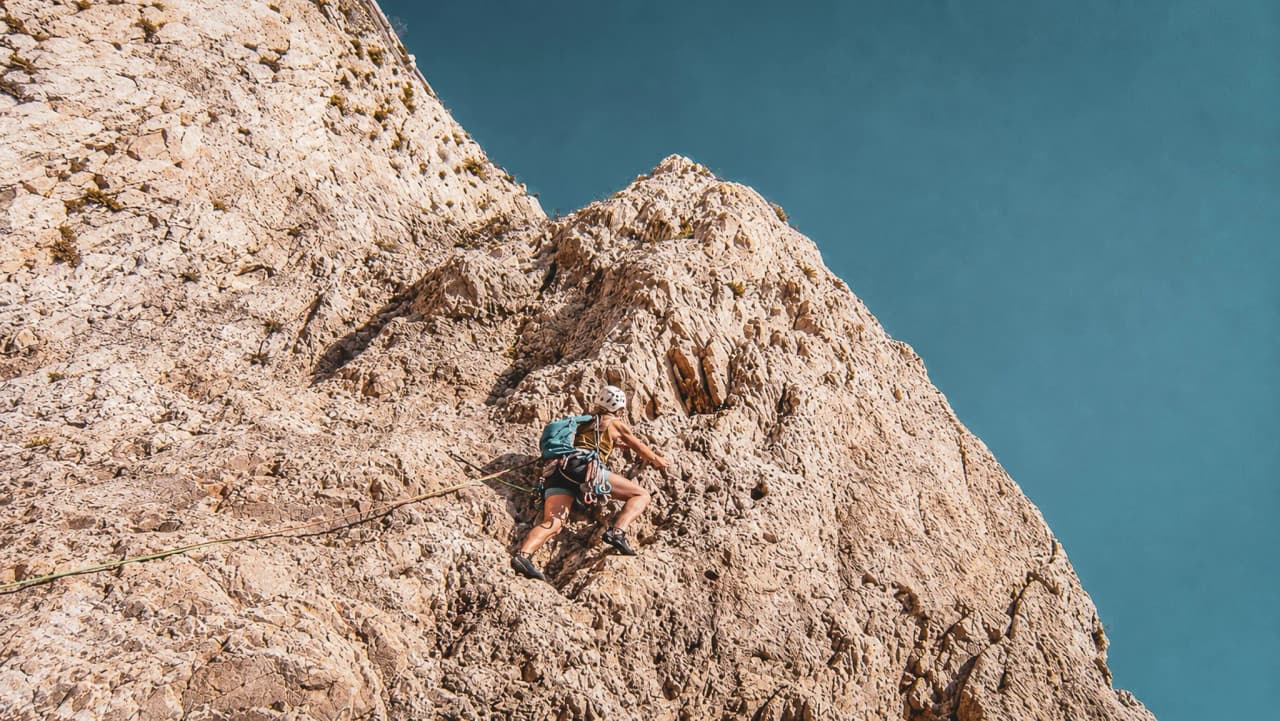 A climber scaling a limestone cliff beneath a blue sky, overlooking the Mediterranean.