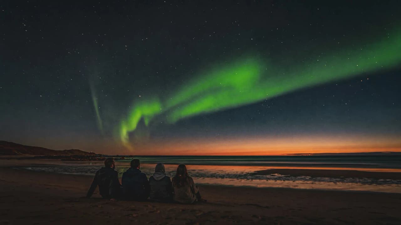 Vier mensen bewonderen het noorderlicht vanaf de kust, onder een sterrenhemel.