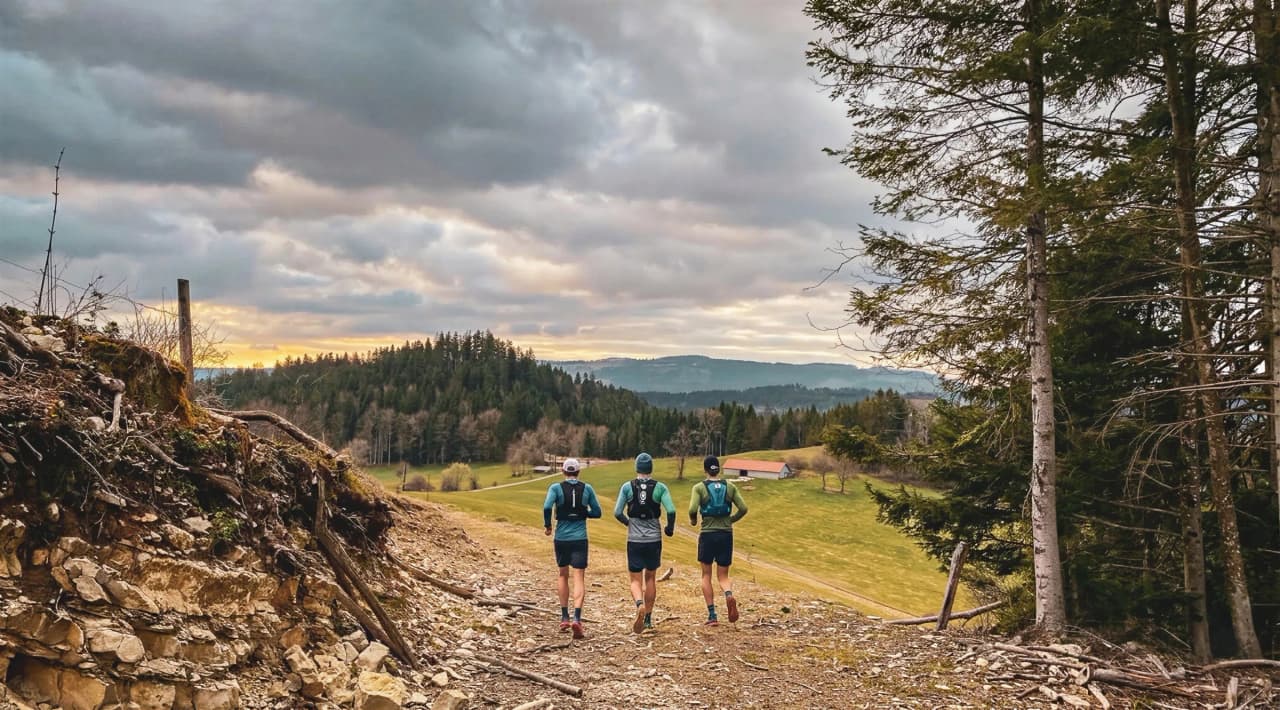 Three runners on a mountain trail, surrounded by lush forests and cloudy skies.