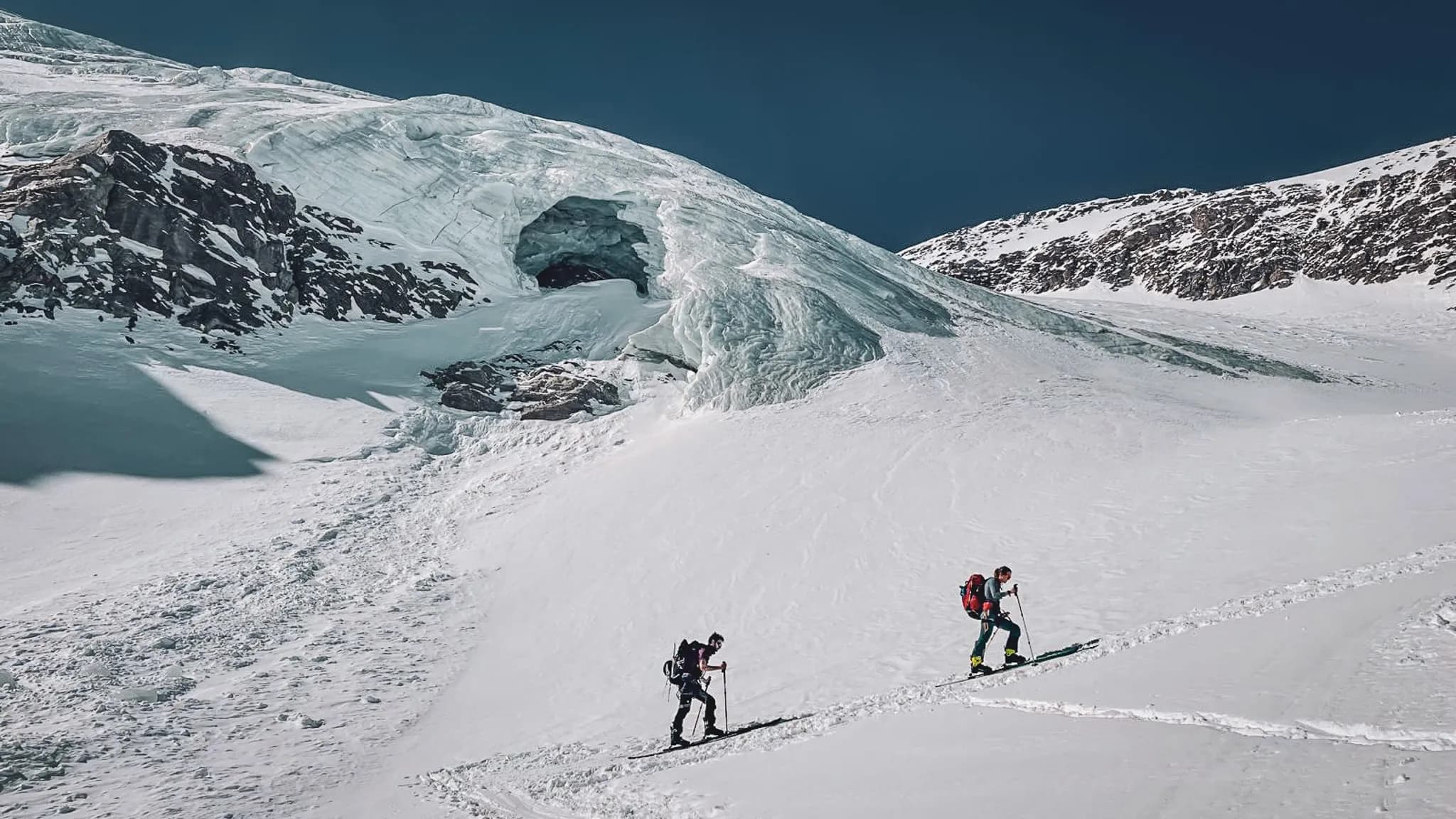 Deux skieurs de randonnée progressent sur un glacier étincelant, entourés de montagnes majestueuses.