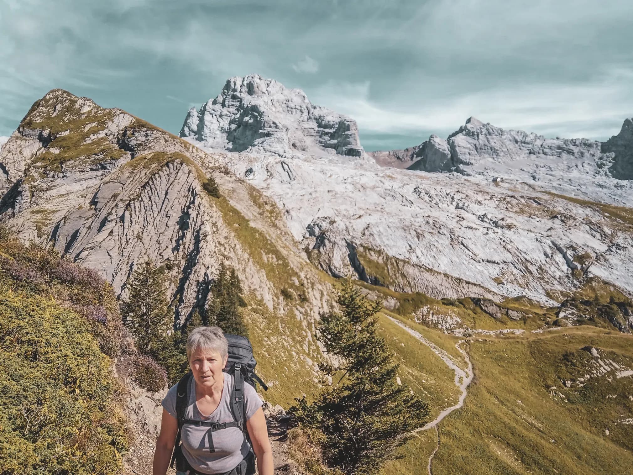 Randonneuse sur un sentier alpin, entourée de montagnes majestueuses et de paysages verdoyants.