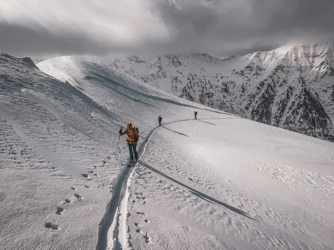 Three skiers set off across a vast snow-covered plateau in the Écrins, beneath a majestic cloudy sky.