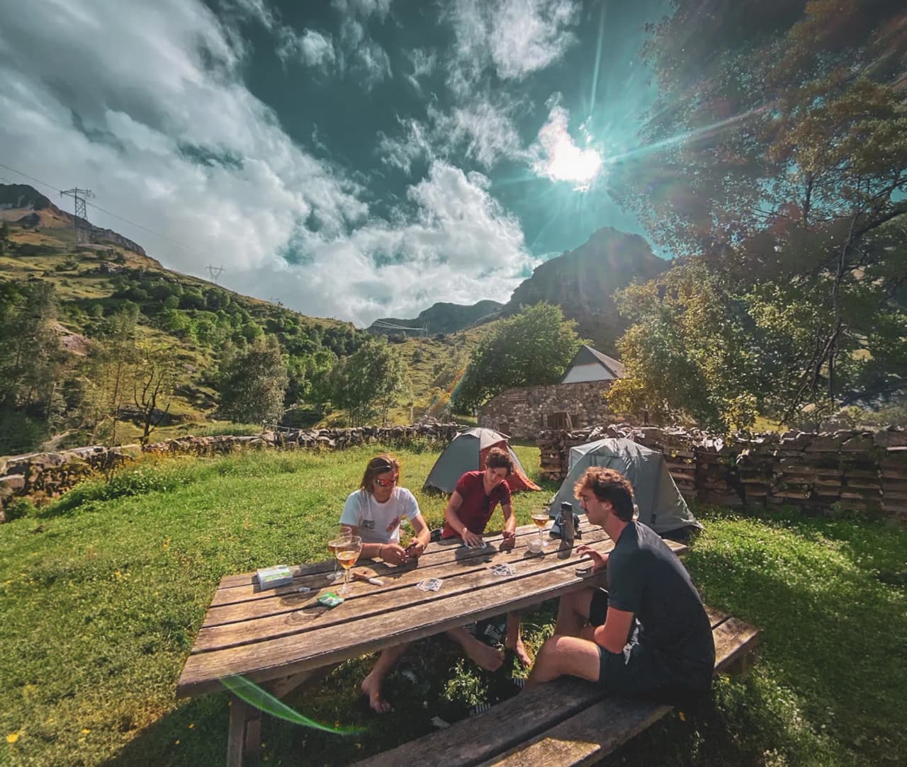 Groupe d'amis autour d'une table en bois, montagne verdoyante en arrière-plan, atmosphère conviviale.