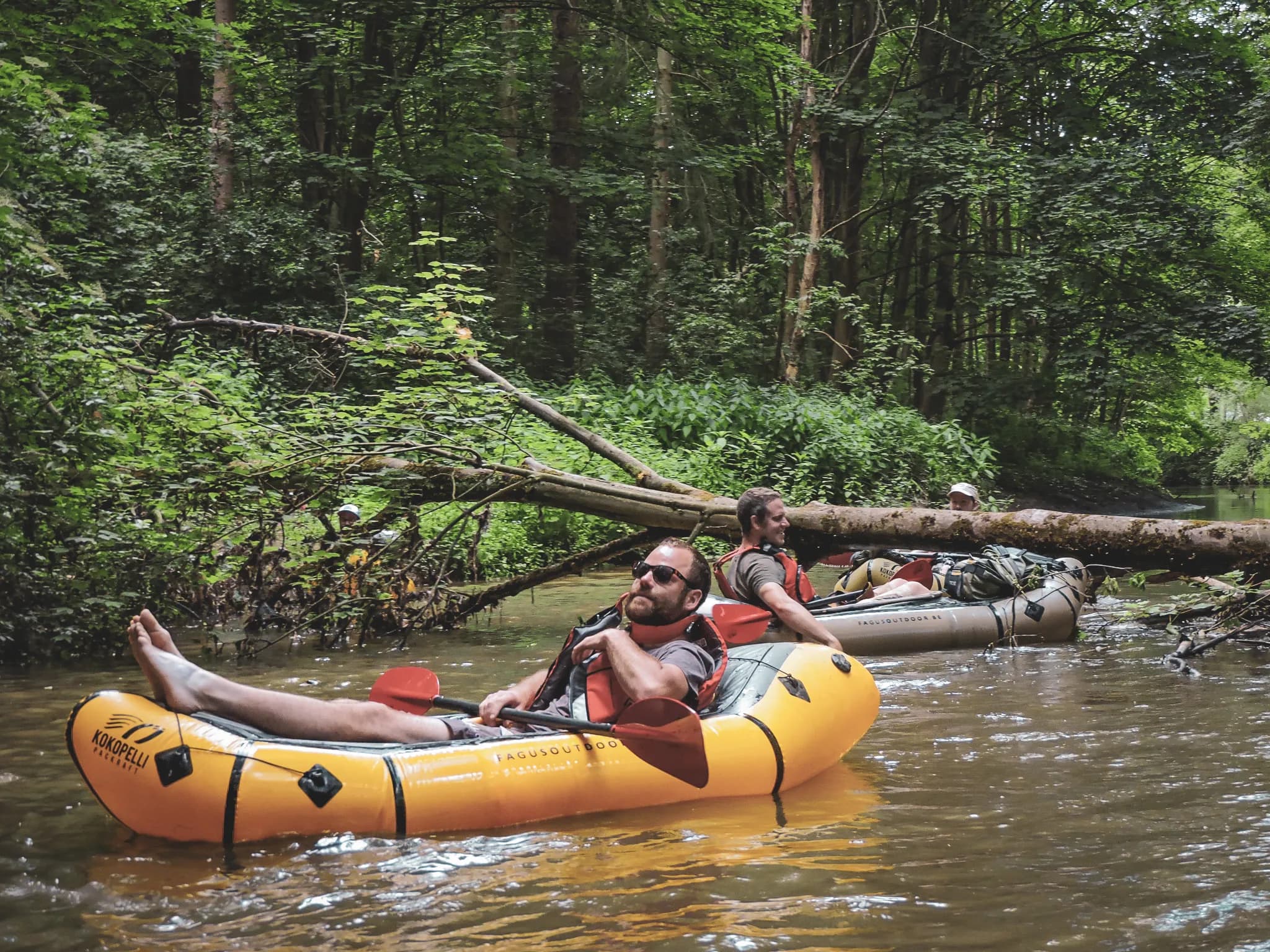 Paddlers relax in a packraft on a green river, surrounded by nature.