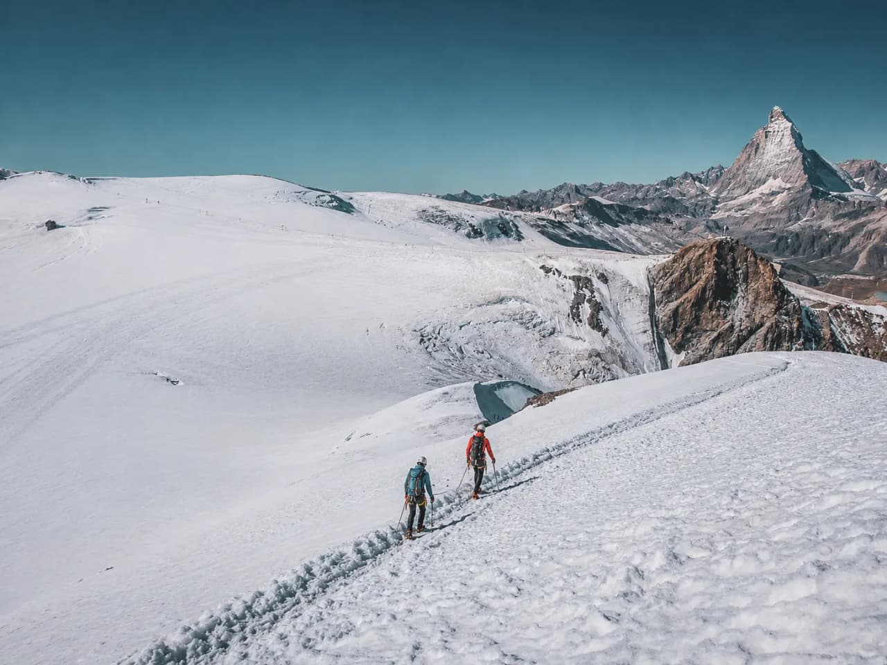 Two mountaineers are making their way across a glacier, with the majestic Breithorn and the Matterhorn in the background.