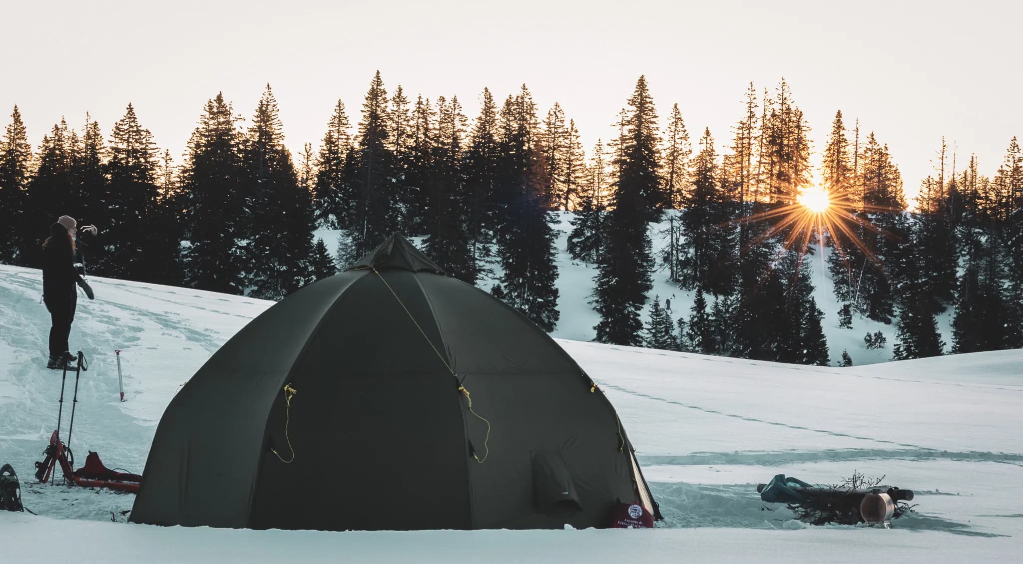 Round tent on the snow, sunrise behind fir trees, peaceful, Nordic atmosphere.