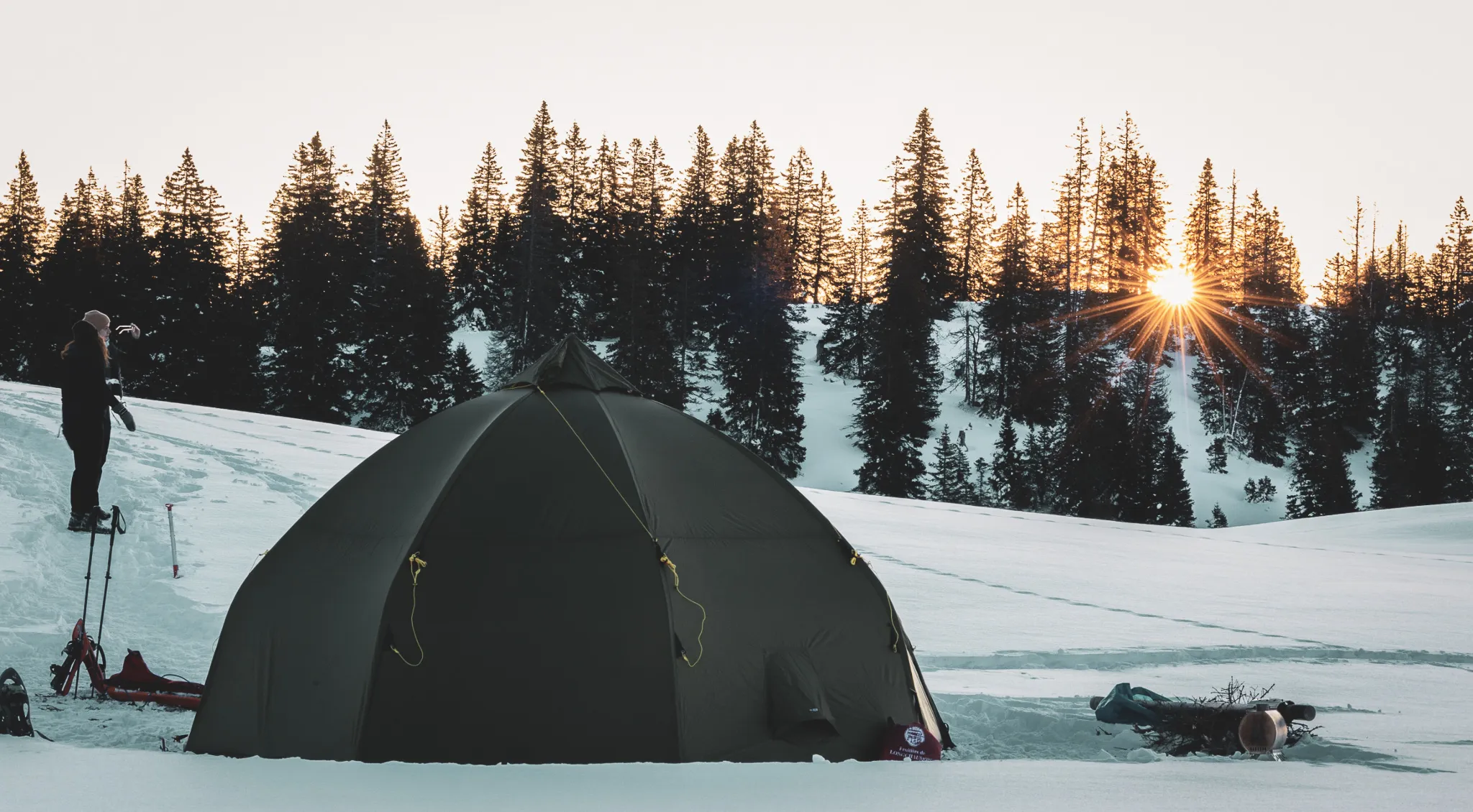 Round tent on the snow, sunrise behind fir trees, peaceful, Nordic atmosphere.