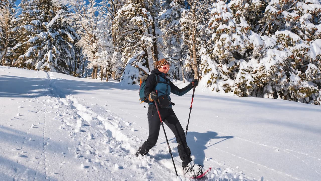 A woman on snowshoes in a snow-covered landscape, surrounded by trees, beneath a brilliant blue sky.