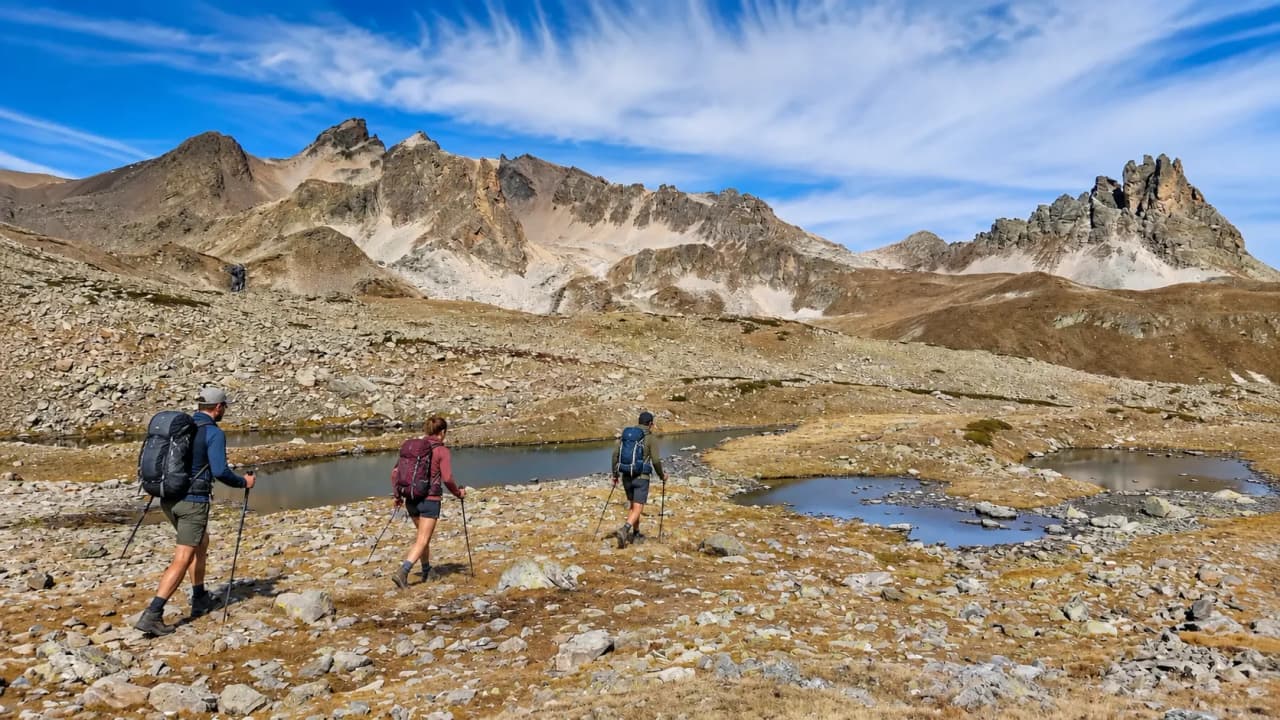 Trois randonneurs traversent un paysage alpin, entourés de montagnes et de lacs scintillants.