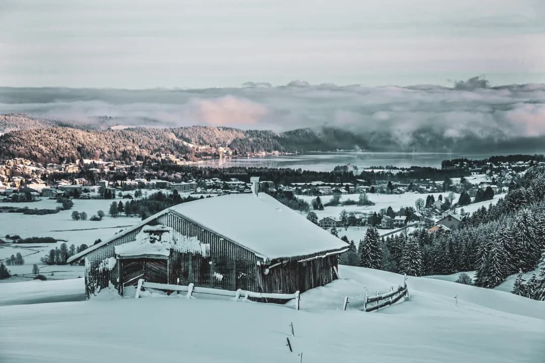 A snowy Jura landscape with a rustic chalet, forests and a tranquil lake in the background.