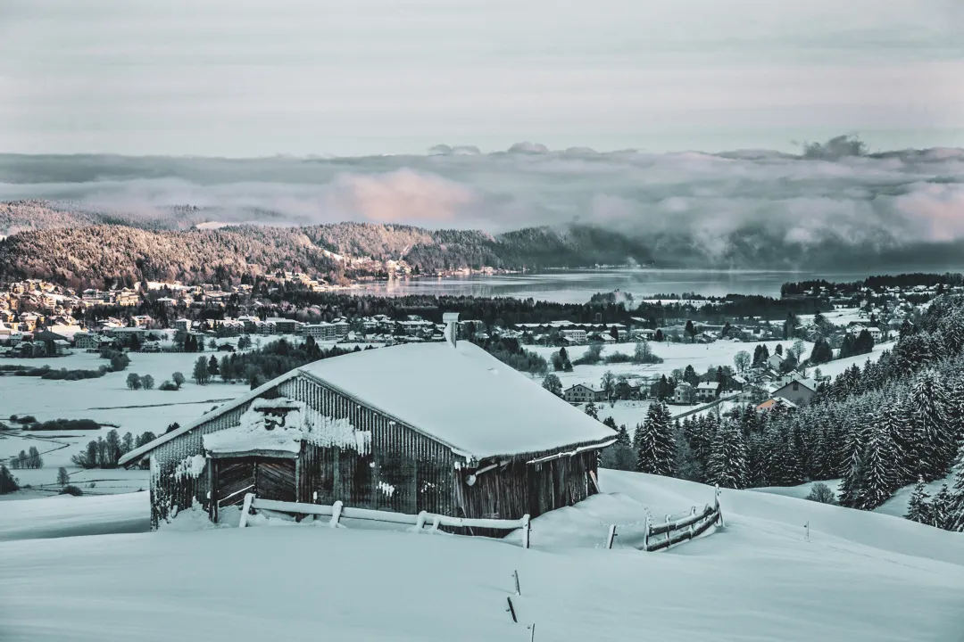 A snowy Jura landscape with a rustic chalet, forests and a tranquil lake in the background.