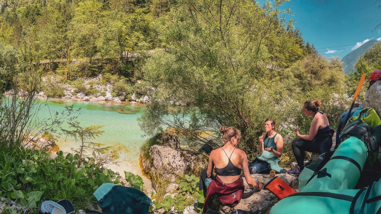 Three adventurous women rest on the banks of a turquoise river surrounded by Alpine greenery.