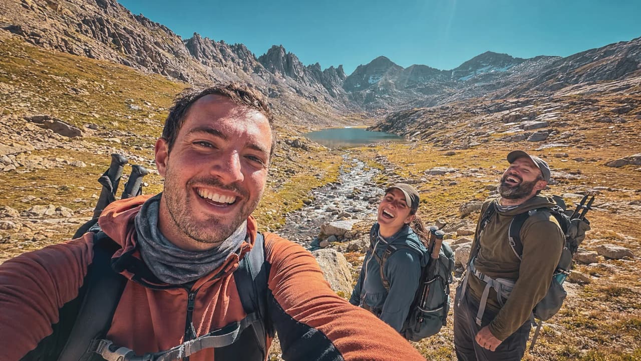 Three smiling hikers in front of an alpine lake and majestic mountains.