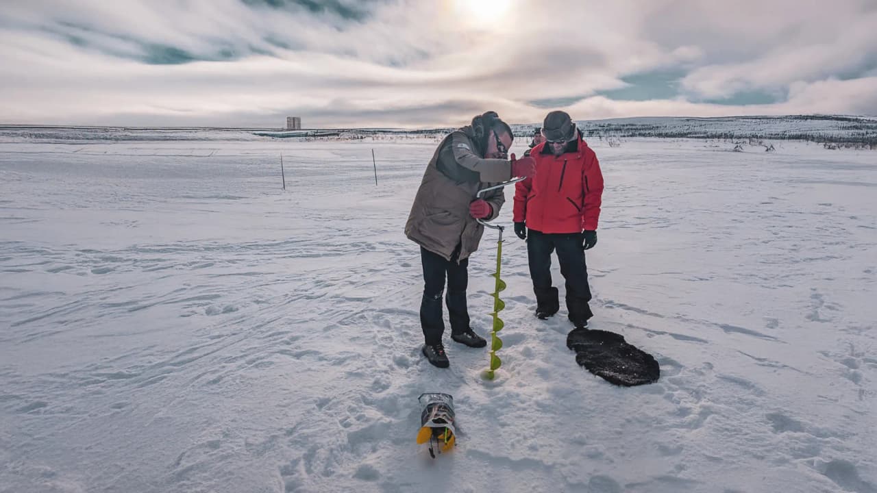 Deux hommes en tenue d’hiver forent un trou dans la neige vierge, découvrant la beauté de la Laponie.