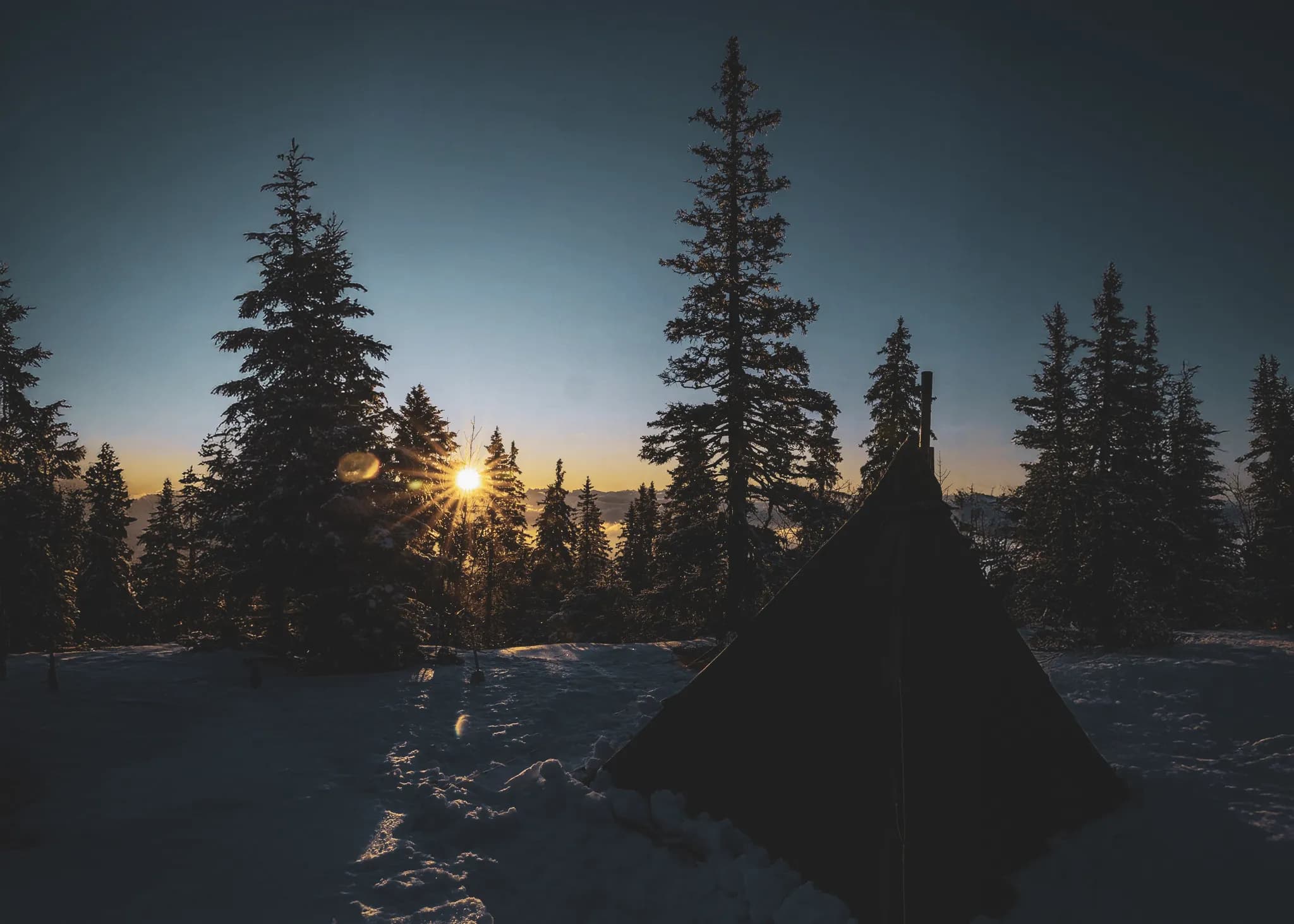 A bright sun rises behind snow-covered fir trees, illuminating a bivouac in the Jura.