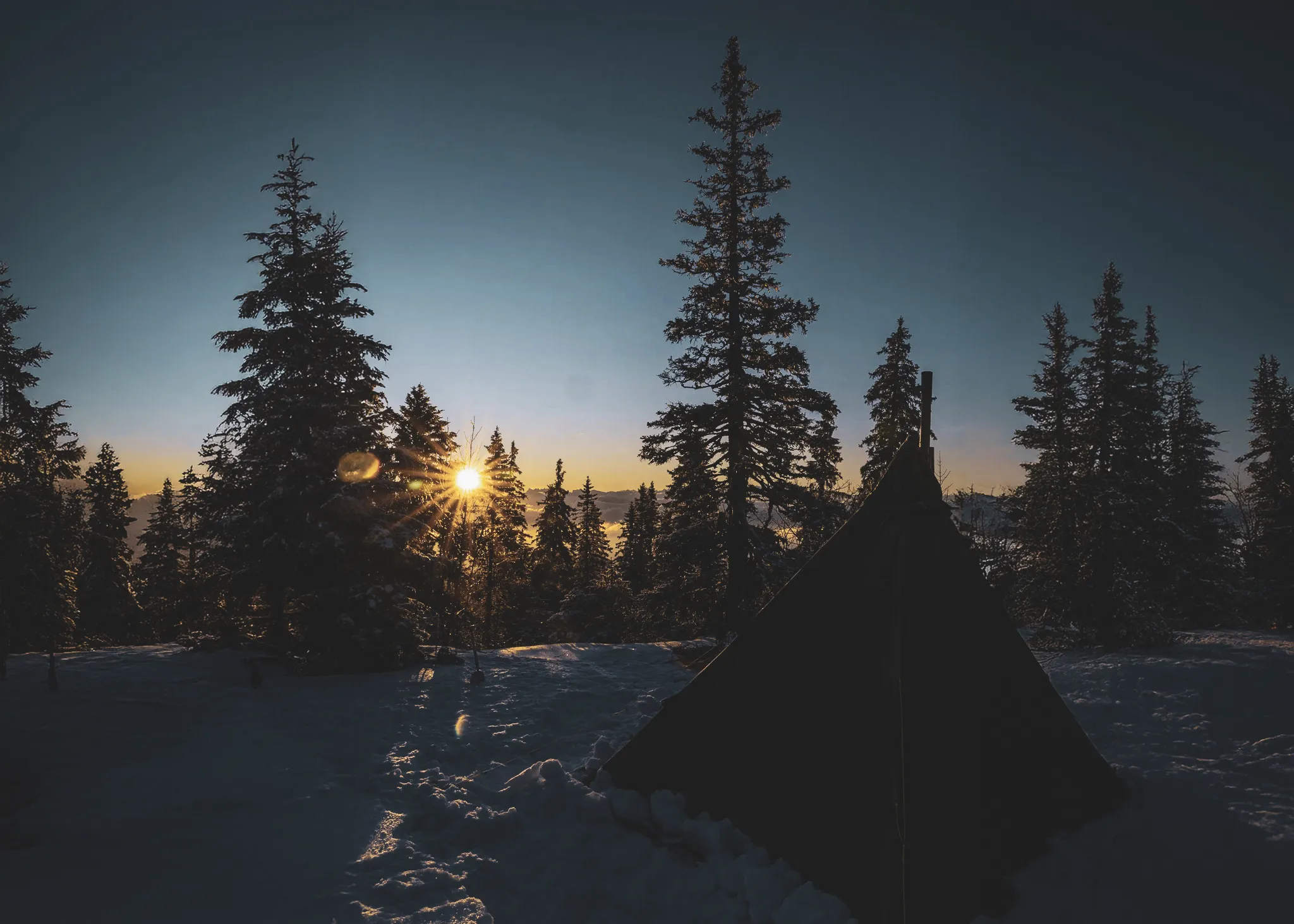 A bright sun rises behind snow-covered fir trees, illuminating a bivouac in the Jura.