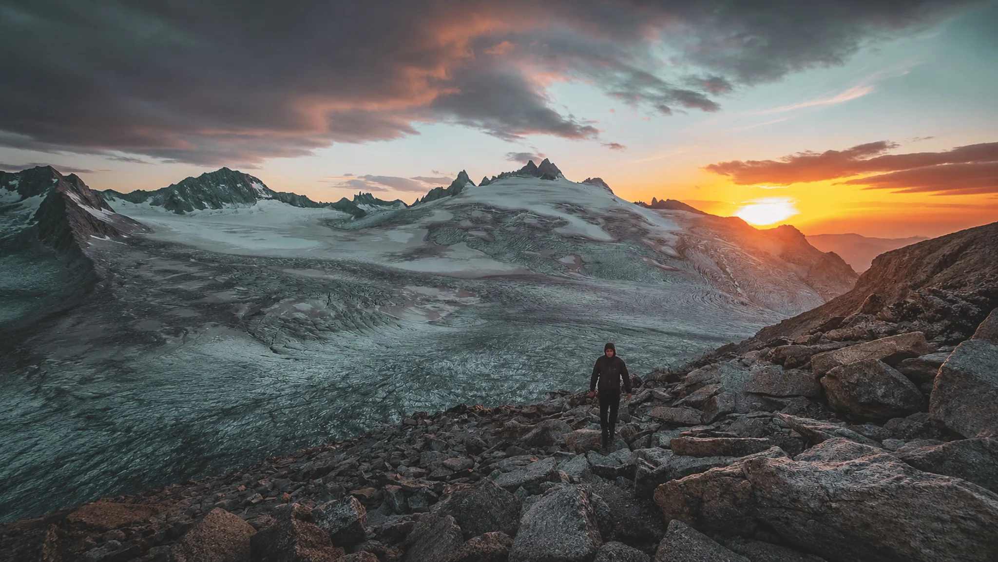 Randonneur sur des rochers face à un glacier au coucher de soleil, ambiance majestueuse.