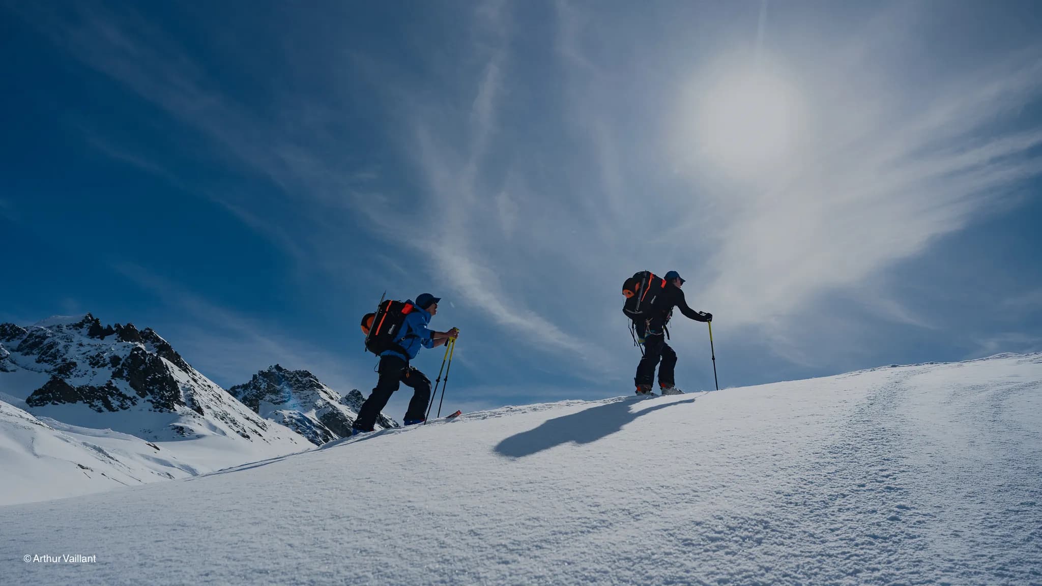 Two skiers climbing a snow-covered ridge under a brilliant blue sky.