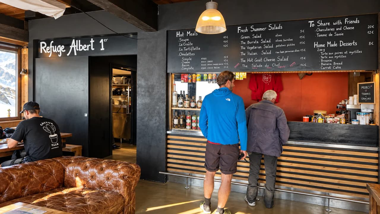 De bar van de berghut Albert 1er, met een krijtbord vol bergmaaltijden en wandelaars aan de toonbank.