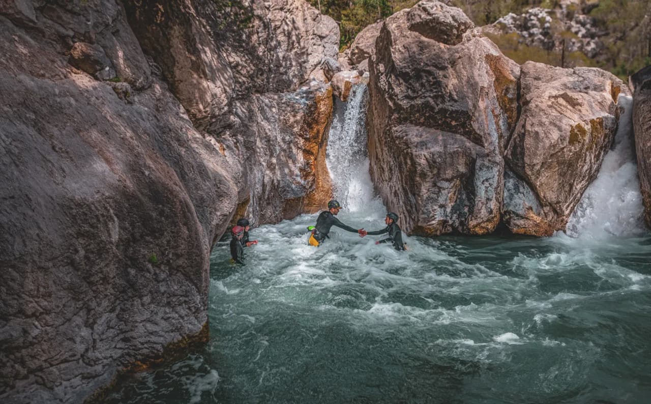 Three adventurers in wetsuits swimming in tumultuous waters between rocks, under a pretty Alpine sky.