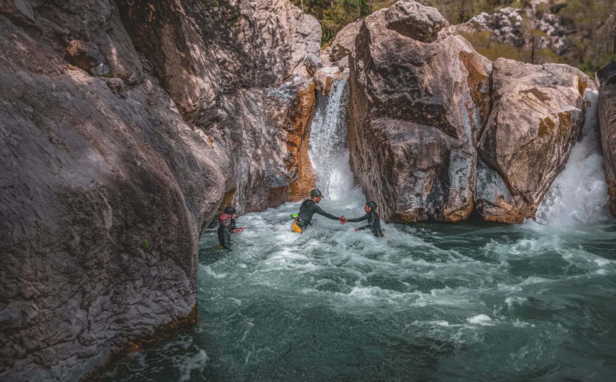 Three adventurers in wetsuits swimming in tumultuous waters between rocks, under a pretty Alpine sky.