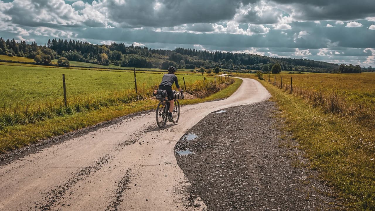 Cycliste sur une route paisible, entouré de champs verdoyants et de forêts au ciel nuageux.