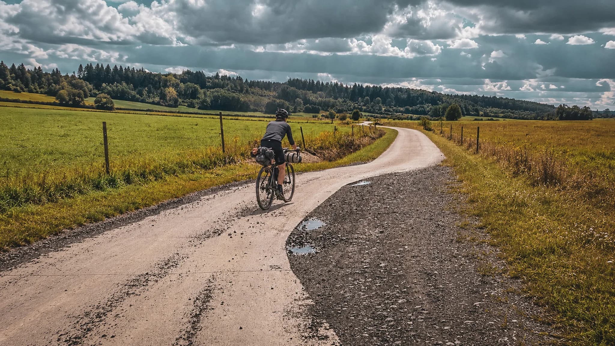 Cycliste sur une route paisible, entouré de champs verdoyants et de forêts au ciel nuageux.
