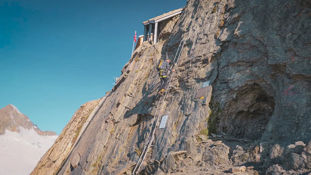 Un alpiniste grimpe une paroi rocheuse vers un refuge en altitude, avec un ciel bleu éclatant.