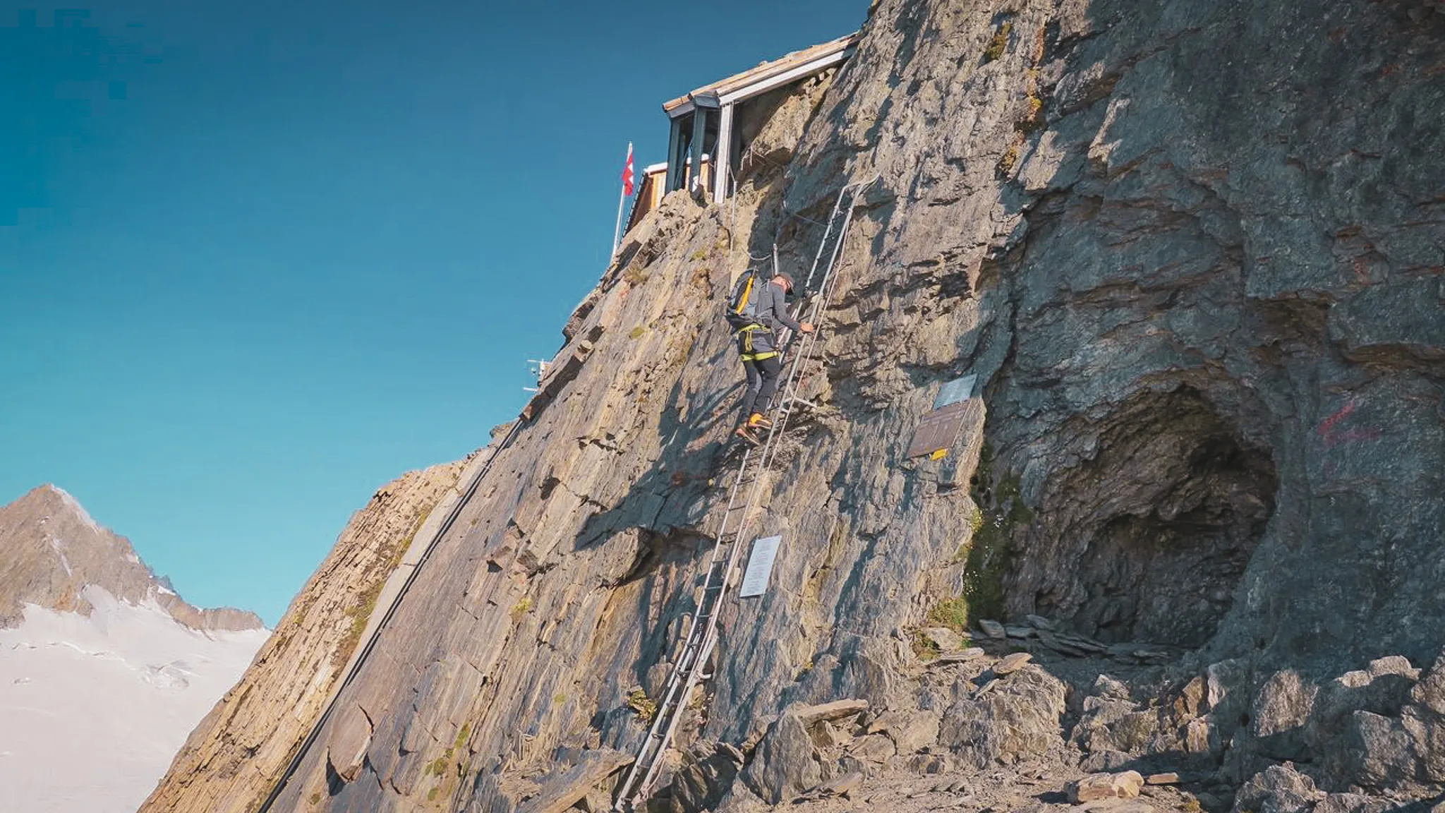 Un alpiniste grimpe une paroi rocheuse vers un refuge en altitude, avec un ciel bleu éclatant.