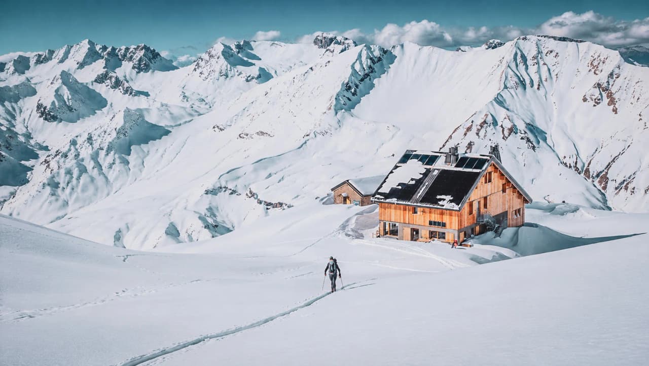 A skier making his way up through the pristine snow towards a wooden mountain hut, surrounded by majestic mountains.