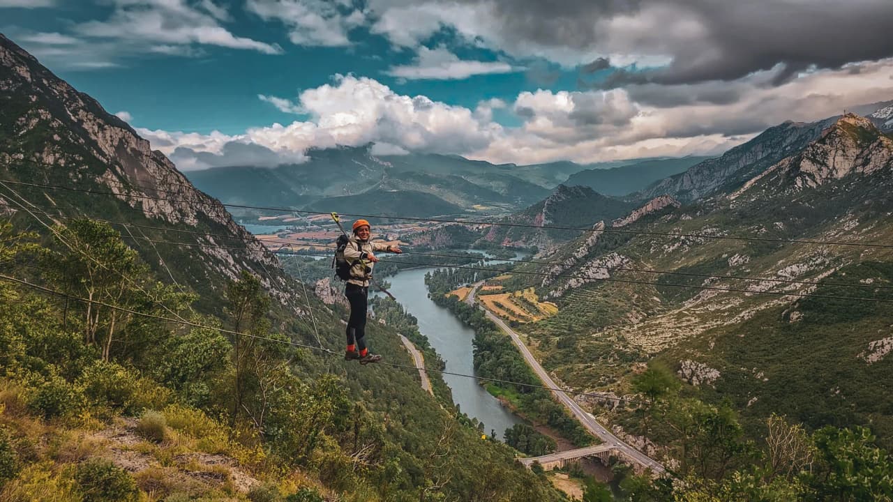 Un aventurier sur une via ferrata surplombant des paysages montagneux et une rivière turquoise.