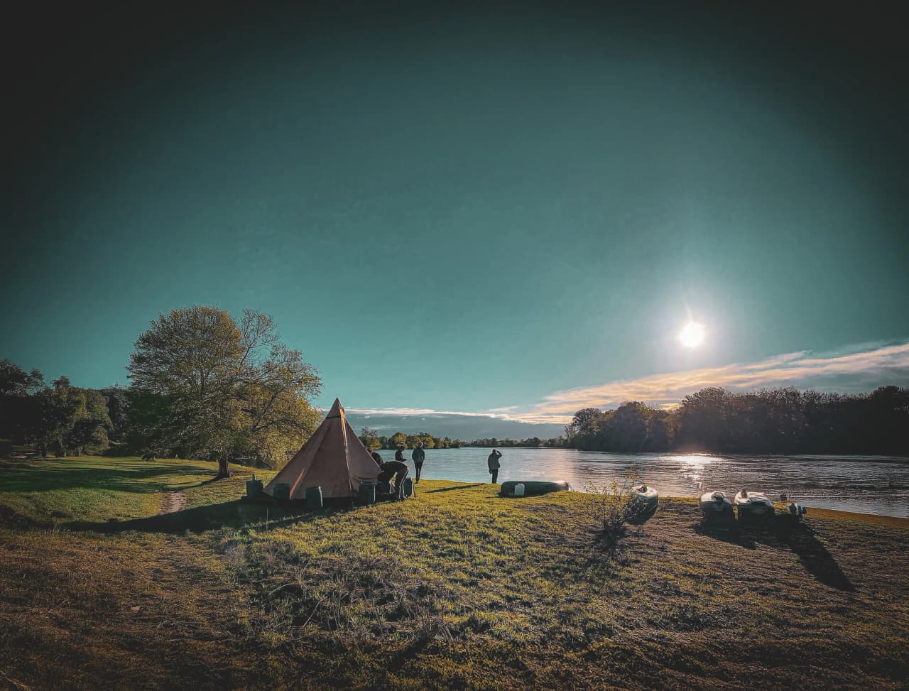 Een rivierlandschap met een strakblauwe lucht en felle zon. Links een grote lommerrijke boom en iets verderop een kegelvormige tent van oranje canvas, omringd door een aantal mensen.
